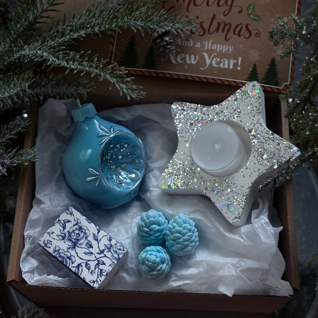 Decorative items including a blue ornament, star-shaped candle holder, and pine cones in a box with Christmas tree branches.
