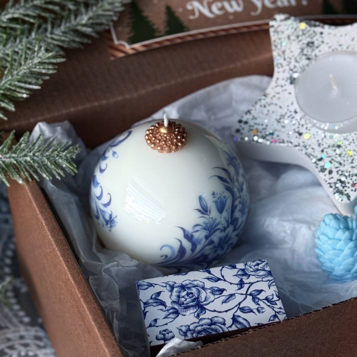 Decorative candle with blue floral design in a gift box with Christmas card and star ornament.