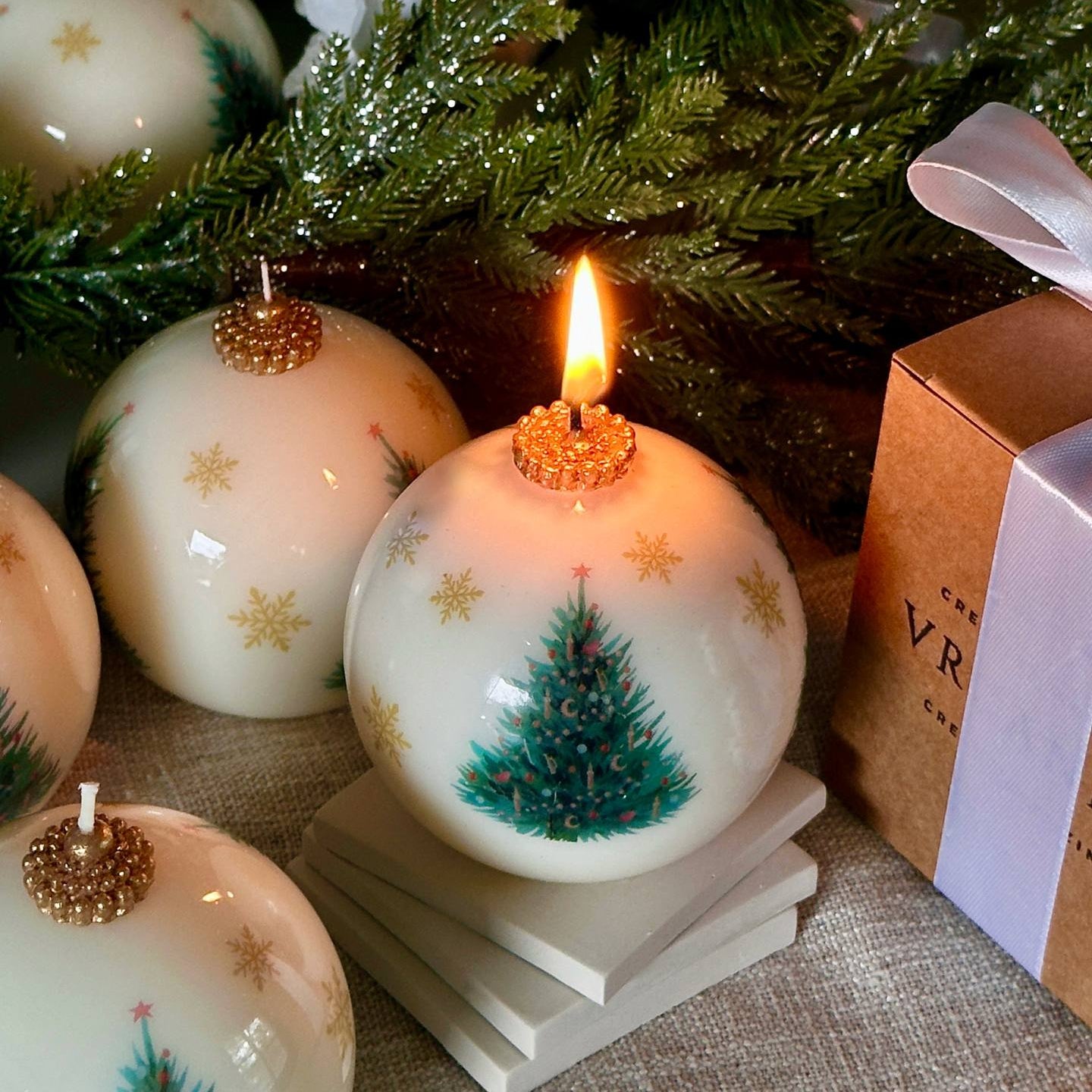 Decorative candles with Christmas tree designs on a table with a tree and gift box in the background.