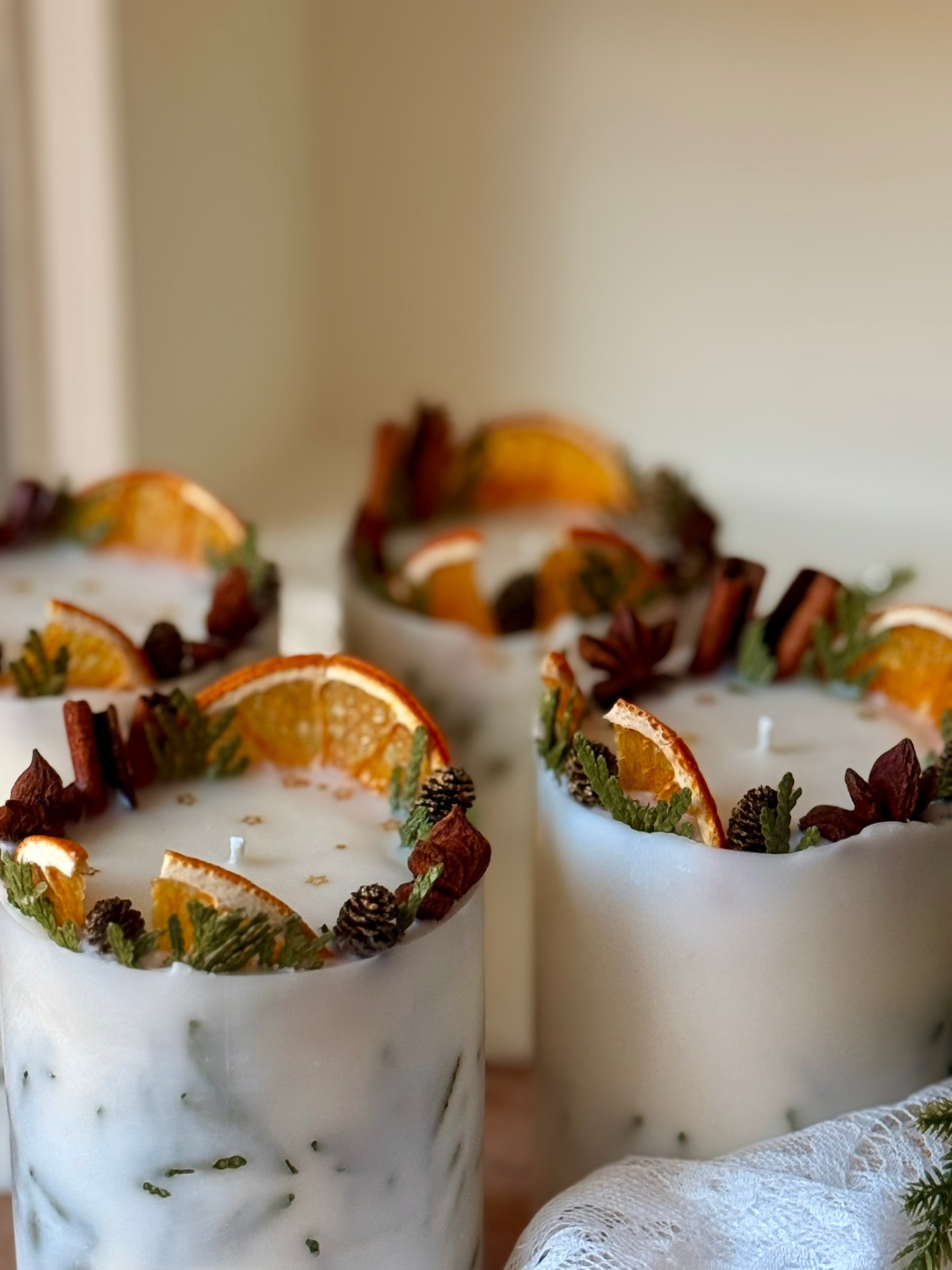 Hand-poured decorative pillar candles topped with dried orange slices, cinnamon sticks, star anise, and cedar greenery on a soft neutral background